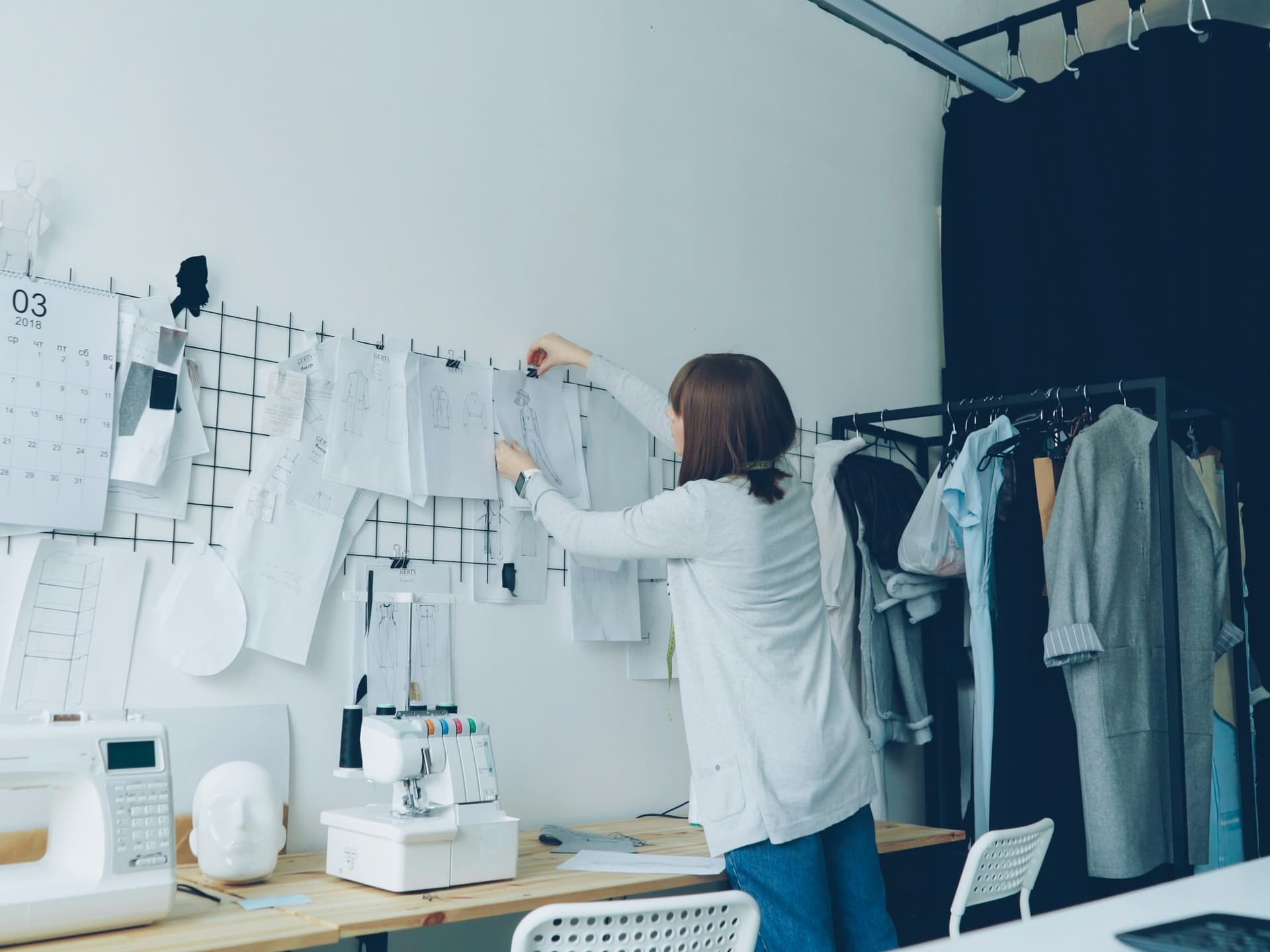 Proprietara unui atelier de modă prinzând schițe pe un panou, între mașinile de cusut și hainele pe umerașe